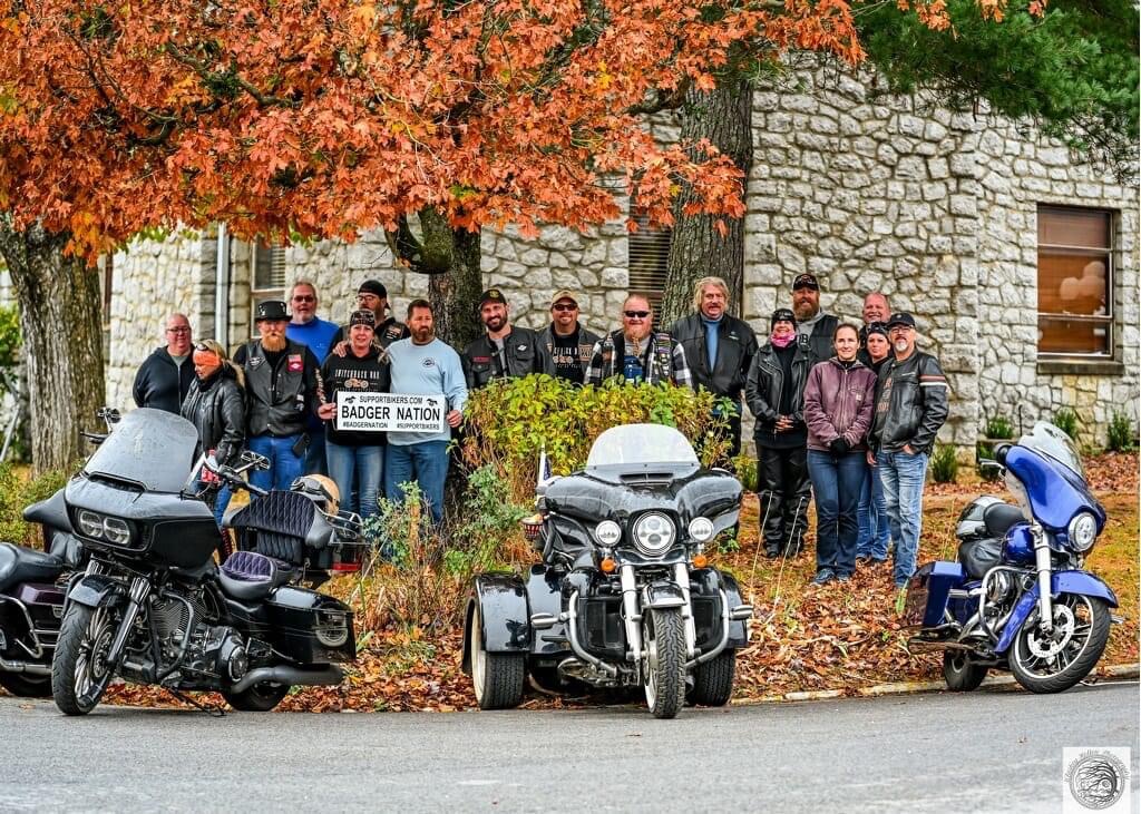 image of motorcyclists hold a banner that says Support Bikers Badger Nation around trees with fall leaves and a motorcycle and trike parked in front of them during an event- Add a Motorcycle Event