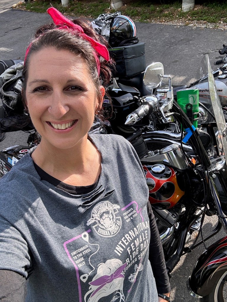 Momma Badger smiling in front of her motorcycle with a hot pink bandana in her hair, showing pride and excitement for women-only riding events.
