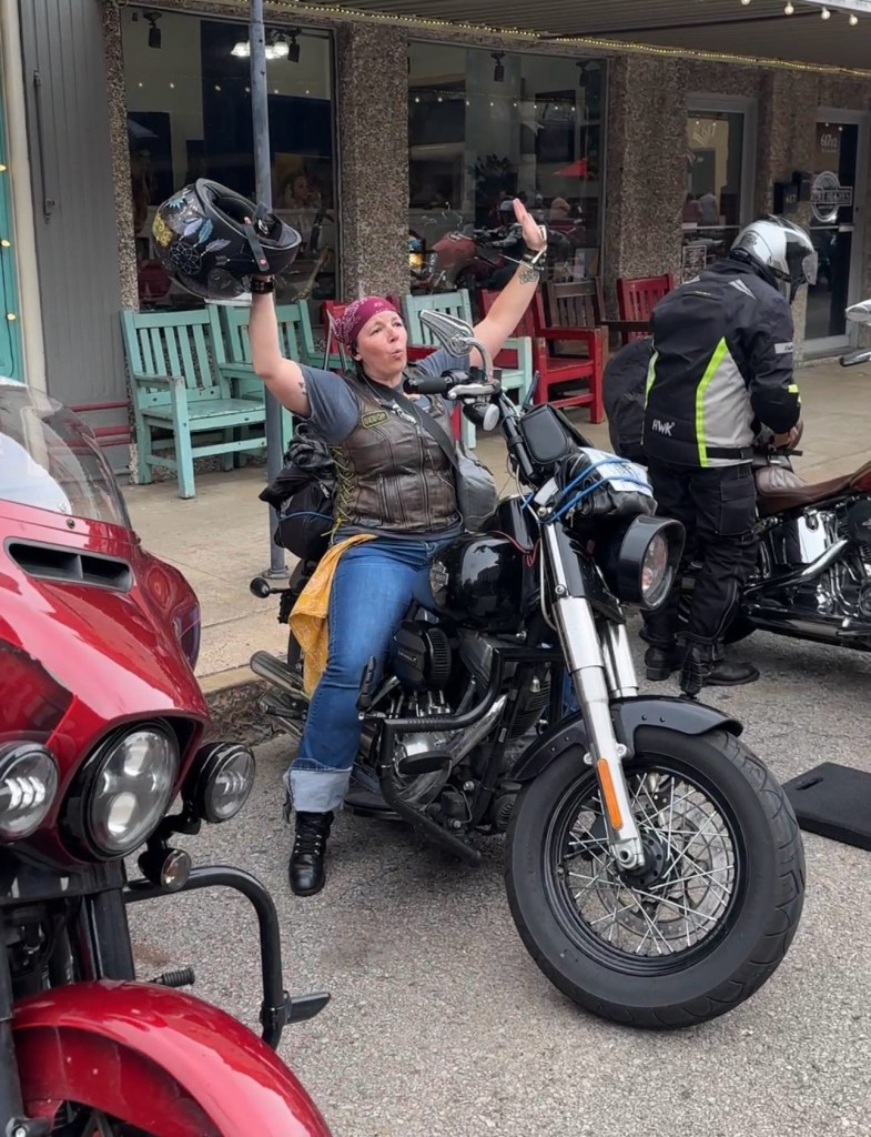 Valerie wearing a helmet and leather jacket, waving while seated on a fully packed touring motorcycle during a long-distance ride through a wooded area.